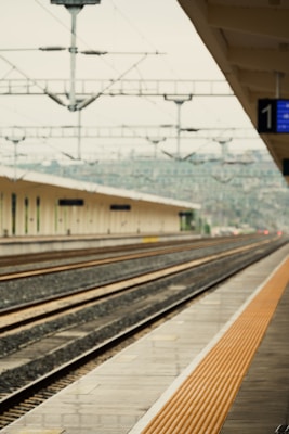 A deserted railway station platform is captured with multiple rail tracks leading into the distance. Overhead, there is an intricate network of electric cables and poles that span across the tracks. The platform is clean and appears well-maintained, with a series of orange safety lines along its edge.