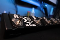 A guitar effects pedal board with multiple foot switches labeled A, B, and C. The surface is metallic with a glossy finish, and logos are present. The background is slightly blurred, creating a studio or music room setting.