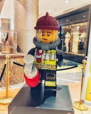 A large LEGO sculpture of a firefighter is prominently displayed in a shopping mall. The figure is wearing a red helmet, a gray suit with a yellow vest, and holding a red and white object. It stands on a platform surrounded by gold and black barriers. The background features a patterned column and a store with mannequins in the window.
