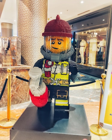 A large LEGO sculpture of a firefighter is prominently displayed in a shopping mall. The figure is wearing a red helmet, a gray suit with a yellow vest, and holding a red and white object. It stands on a platform surrounded by gold and black barriers. The background features a patterned column and a store with mannequins in the window.