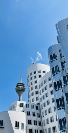 A modern cityscape featuring uniquely designed, curving buildings with reflective surfaces and numerous windows. A prominent telecommunications tower rises in the background, with a blue sky and a few clouds visible above.