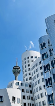 A modern cityscape featuring uniquely designed, curving buildings with reflective surfaces and numerous windows. A prominent telecommunications tower rises in the background, with a blue sky and a few clouds visible above.