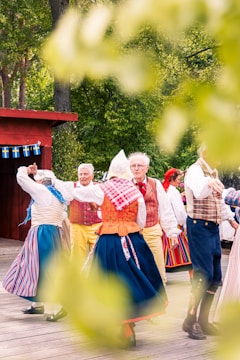 A group of people dressed in traditional folk costumes are dancing outdoors. The scene includes vibrant clothing with stripes and checks in colors like red, blue, and yellow. The background shows lush green trees, and small flags are hung on a wooden structure, suggesting a cultural or national celebration.