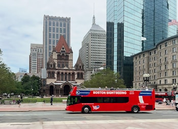 A red sightseeing bus is parked in front of a historic church with ornate architecture and large windows. Modern skyscrapers and classic buildings surround the area, with green trees scattered throughout the park, where people are walking and sitting.