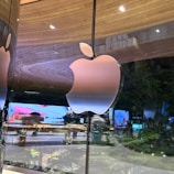 A large, shiny Apple logo is prominently displayed on a reflective glass surface, with the background showing a tree-lined street and illuminated advertisements. The indoor setting captures reflections which create a sophisticated and modern ambiance.