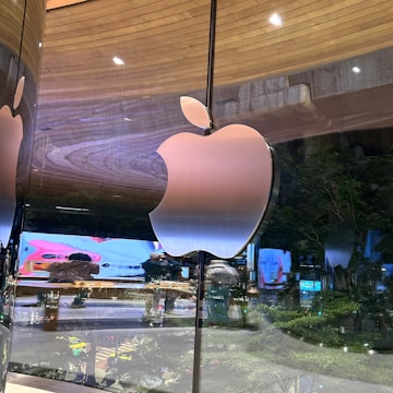 A large, shiny Apple logo is prominently displayed on a reflective glass surface, with the background showing a tree-lined street and illuminated advertisements. The indoor setting captures reflections which create a sophisticated and modern ambiance.