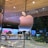 A large, shiny Apple logo is prominently displayed on a reflective glass surface, with the background showing a tree-lined street and illuminated advertisements. The indoor setting captures reflections which create a sophisticated and modern ambiance.