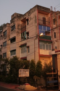 A multi-story residential building exhibits signs of aging with visible weathering and faded paint. Several windows are adorned with varied grill designs, and a few are covered with blue and green panels. Electrical wires are prominently visible running across the scene. In the foreground, a sign in a non-English script, likely Hindi, stands beside well-maintained vegetation. The image is captured at dusk, with a streetlight casting a warm glow.