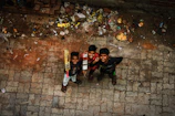 Three boys are looking upward while standing on a rough, stone-paved ground next to some scattered trash and debris. One of the boys holds a cricket bat, suggesting they might be playing a game. The boys appear cheerful.
