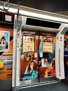 A subway train interior features vibrant and artistic advertisements on the doors. The artwork includes a man wearing a hat and a woman with curly hair, both depicted in a colorful, modern style. The surrounding walls are adorned with abstract designs and promotional posters.