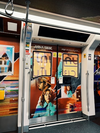 A subway train interior features vibrant and artistic advertisements on the doors. The artwork includes a man wearing a hat and a woman with curly hair, both depicted in a colorful, modern style. The surrounding walls are adorned with abstract designs and promotional posters.