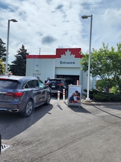 A line of cars is waiting at the entrance of a building with a red and white facade featuring a maple leaf symbol. The area is well-lit with street lamps and surrounded by trees and a clear sky with some clouds. There is a promotional sign with an image of two people next to the entrance, and white and orange traffic cones are positioned around the entrance.