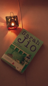 A candle holder shaped like a small house with a lit candle inside casts a warm glow on a nearby book. The book title is 'sarah Jio', showing a cover with an image of a bed with pillows and decorative elements.