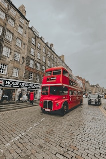 A classic red double-decker bus drives down a cobblestone street lined with historic buildings. The buildings have a traditional architectural style with multiple windows and shops on the ground floor, including one with a sign that reads 'House of Scotland'. The sky is overcast, adding a muted tone to the scene.