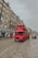A classic red double-decker bus drives down a cobblestone street lined with historic buildings. The buildings have a traditional architectural style with multiple windows and shops on the ground floor, including one with a sign that reads 'House of Scotland'. The sky is overcast, adding a muted tone to the scene.