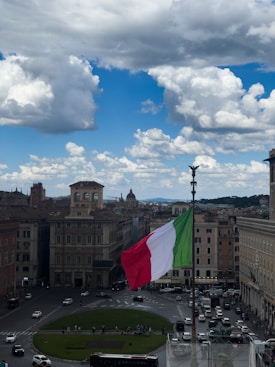 A large Italian flag waves prominently over a bustling city square, surrounded by historic buildings under a partly cloudy sky. The atmosphere is lively with people and vehicles moving around the circular road. Classical architecture dominates the scene, with a backdrop of distant hills visible under the expansive sky.