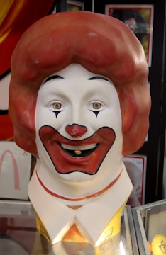 A large sculpture of a clown head with exaggerated features, including a big red nose, wide red lips, and a large red wig. The clown has a prominent smile and is wearing a yellow and white outfit. The background consists of various images or posters.