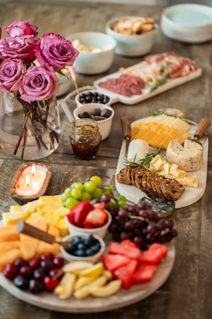 A beautifully arranged charcuterie board with an assortment of fruits, cheeses, and meats is displayed on a wooden table. The scene is accented with a vase of vibrant pink roses and the warm light of a lit candle. Various bowls contain olives, nuts, and small slices of bread, adding to the abundance and variety of the spread.