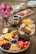 A beautifully arranged charcuterie board with an assortment of fruits, cheeses, and meats is displayed on a wooden table. The scene is accented with a vase of vibrant pink roses and the warm light of a lit candle. Various bowls contain olives, nuts, and small slices of bread, adding to the abundance and variety of the spread.