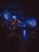 A dimly lit bar with a moody ambiance. The warm glow of hanging lights contrasts with neon blue signage. Patrons are seated, creating a cozy and intimate atmosphere. The space features a bar counter, tables, and stools.