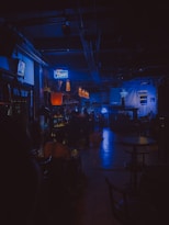 A dimly lit bar with a moody ambiance. The warm glow of hanging lights contrasts with neon blue signage. Patrons are seated, creating a cozy and intimate atmosphere. The space features a bar counter, tables, and stools.