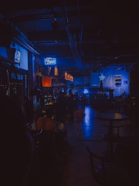 A dimly lit bar with a moody ambiance. The warm glow of hanging lights contrasts with neon blue signage. Patrons are seated, creating a cozy and intimate atmosphere. The space features a bar counter, tables, and stools.