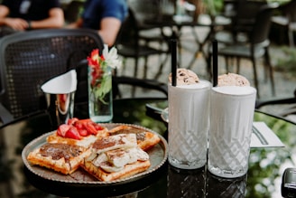 A glass table is set with a plate of waffles topped with chocolate spread and fresh strawberries, along with two tall milkshakes garnished with cookies. A small vase with flowers is placed nearby, adding a touch of color to the serene outdoor café setting.