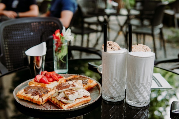 A glass table is set with a plate of waffles topped with chocolate spread and fresh strawberries, along with two tall milkshakes garnished with cookies. A small vase with flowers is placed nearby, adding a touch of color to the serene outdoor café setting.