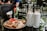 A glass table is set with a plate of waffles topped with chocolate spread and fresh strawberries, along with two tall milkshakes garnished with cookies. A small vase with flowers is placed nearby, adding a touch of color to the serene outdoor café setting.