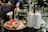 A glass table is set with a plate of waffles topped with chocolate spread and fresh strawberries, along with two tall milkshakes garnished with cookies. A small vase with flowers is placed nearby, adding a touch of color to the serene outdoor café setting.