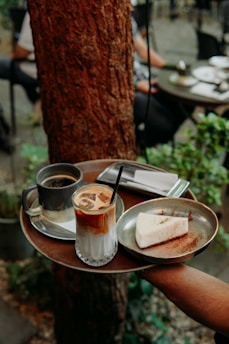 A hand holds a tray with a glass of iced coffee, a slice of cake with a dusting of cocoa, and a cup of hot coffee. The background features a rustic outdoor café setting with wooden tables and greenery.