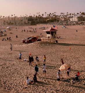 A sandy beach with several people gathered around, engaged in leisure activities. Some individuals are standing and conversing, while others are sitting on the sand under a colorful umbrella. In the background, there is a lifeguard tower with an American flag, a couple of vehicles, and more people spread out across the beach. Tall palm trees line the horizon, with buildings and parked cars visible further back.
