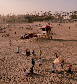 A sandy beach with several people gathered around, engaged in leisure activities. Some individuals are standing and conversing, while others are sitting on the sand under a colorful umbrella. In the background, there is a lifeguard tower with an American flag, a couple of vehicles, and more people spread out across the beach. Tall palm trees line the horizon, with buildings and parked cars visible further back.