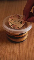 A smiling woman packaging her homemade marmitas in simple, clear containers at her kitchen table.