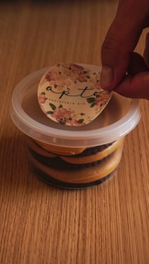 A hand is peeling off a floral sticker labeled 'apto' from the top of a transparent plastic container filled with layered brown and black dessert. The container is placed on a wooden surface.