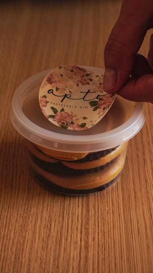 A hand is peeling off a floral sticker labeled 'apto' from the top of a transparent plastic container filled with layered brown and black dessert. The container is placed on a wooden surface.
