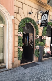 A storefront adorned with green garlands and pink flowers framing the entrance of a jewelry shop. The stone facade features detailed masonry, and there's a sign displaying 'V. Vittoria Jewels'. The entrance door is open, revealing part of the interior, and a large potted plant is positioned nearby.