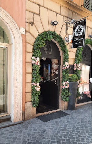 A storefront adorned with green garlands and pink flowers framing the entrance of a jewelry shop. The stone facade features detailed masonry, and there's a sign displaying 'V. Vittoria Jewels'. The entrance door is open, revealing part of the interior, and a large potted plant is positioned nearby.