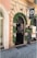 A storefront adorned with green garlands and pink flowers framing the entrance of a jewelry shop. The stone facade features detailed masonry, and there's a sign displaying 'V. Vittoria Jewels'. The entrance door is open, revealing part of the interior, and a large potted plant is positioned nearby.