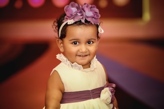 A young child with curly hair is wearing a sleeveless white dress with a purple sash and flower detail. A purple floral headband decorates the child’s head. The background is softly blurred with warm hues of brown and purple.
