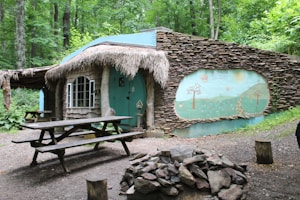 A whimsical, rustic cabin nestled in a forest setting. The cabin features a stone facade with a straw thatched roof, a vibrant teal door, and a unique mural depicting trees and a winding path. Picnic tables and logs are situated outside, with a pile of rocks nearby. Lush green trees surround the area, creating a serene woodland atmosphere.