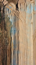 The image shows an aerial view of a landscape with various tire tracks imprinted in the sand. The terrain features both brown earthy and green patches, along with blue-tinted sections. The alignment of tire tracks suggests heavy machinery movement or construction activities.