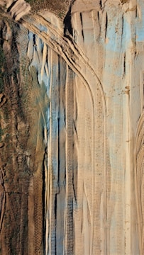 The image shows an aerial view of a landscape with various tire tracks imprinted in the sand. The terrain features both brown earthy and green patches, along with blue-tinted sections. The alignment of tire tracks suggests heavy machinery movement or construction activities.