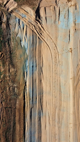 The image shows an aerial view of a landscape with various tire tracks imprinted in the sand. The terrain features both brown earthy and green patches, along with blue-tinted sections. The alignment of tire tracks suggests heavy machinery movement or construction activities.