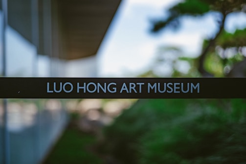 A sign with the text 'LUO HONG ART MUSEUM' is centered in the foreground, with a modern architectural structure and blurred greenery in the background.