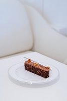A rectangular chocolate dessert on a white plate, featuring detailed topping work with small pieces of nut and a decorative chocolate stick. The background consists of a cream-colored couch and table, providing a minimal and elegant setting.