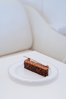 A rectangular chocolate dessert on a white plate, featuring detailed topping work with small pieces of nut and a decorative chocolate stick. The background consists of a cream-colored couch and table, providing a minimal and elegant setting.