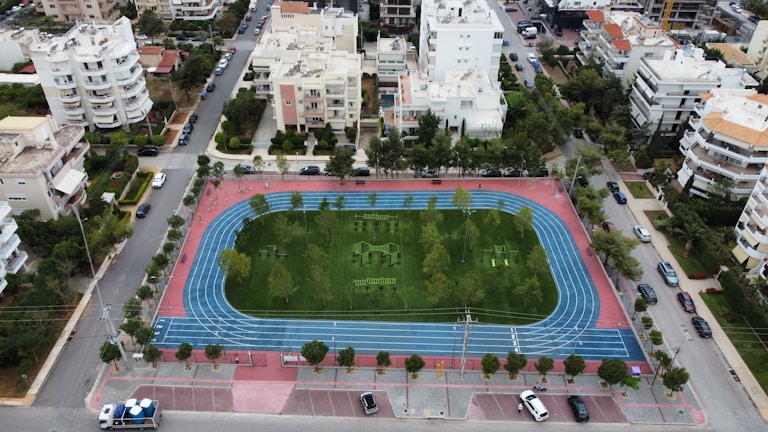 An aerial view of a sports complex featuring a blue running track surrounding a green field with workout equipment and trees. The area is bordered by residential buildings and streets with parked cars and greenery.