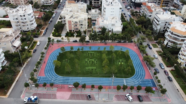 An aerial view of a sports complex featuring a blue running track surrounding a green field with workout equipment and trees. The area is bordered by residential buildings and streets with parked cars and greenery.