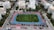 An aerial view of a sports complex featuring a blue running track surrounding a green field with workout equipment and trees. The area is bordered by residential buildings and streets with parked cars and greenery.